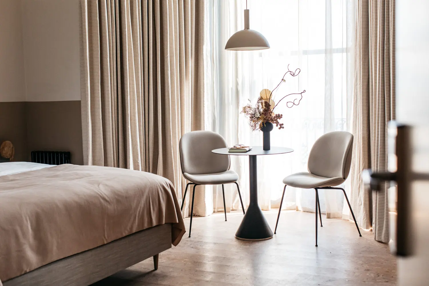 A hotel bedroom featuring a bed with a throw, full length curtains, a small table and two chairs, and a vase of fresh flowers