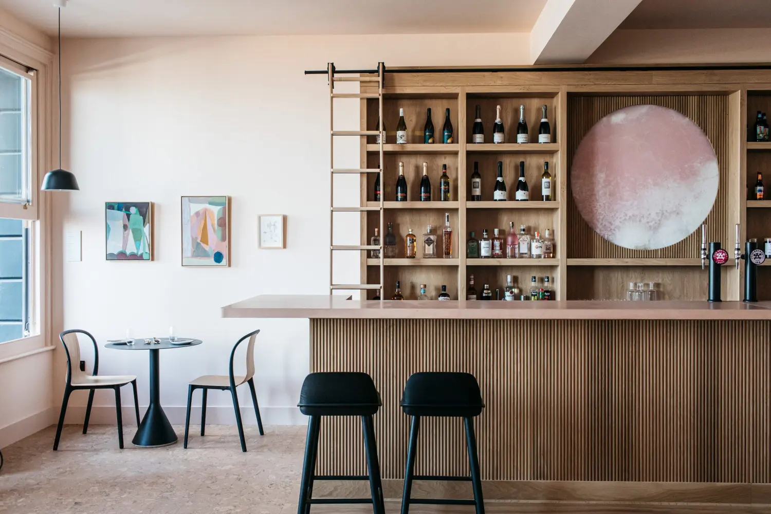 A wood-panelled bar with bar stools, a small table and two matching chairs, and pastel-coloured artworks