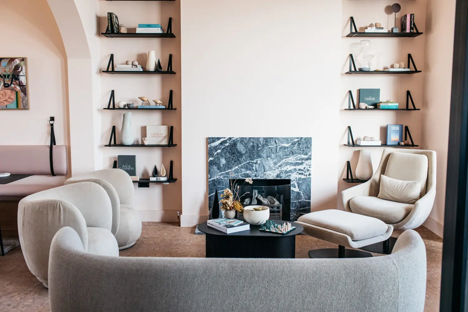 A liunge area of a bar featuring a marble fireplace framed bit two allcoved shelving units, and surrounding by curving armchairs and a matching sofa